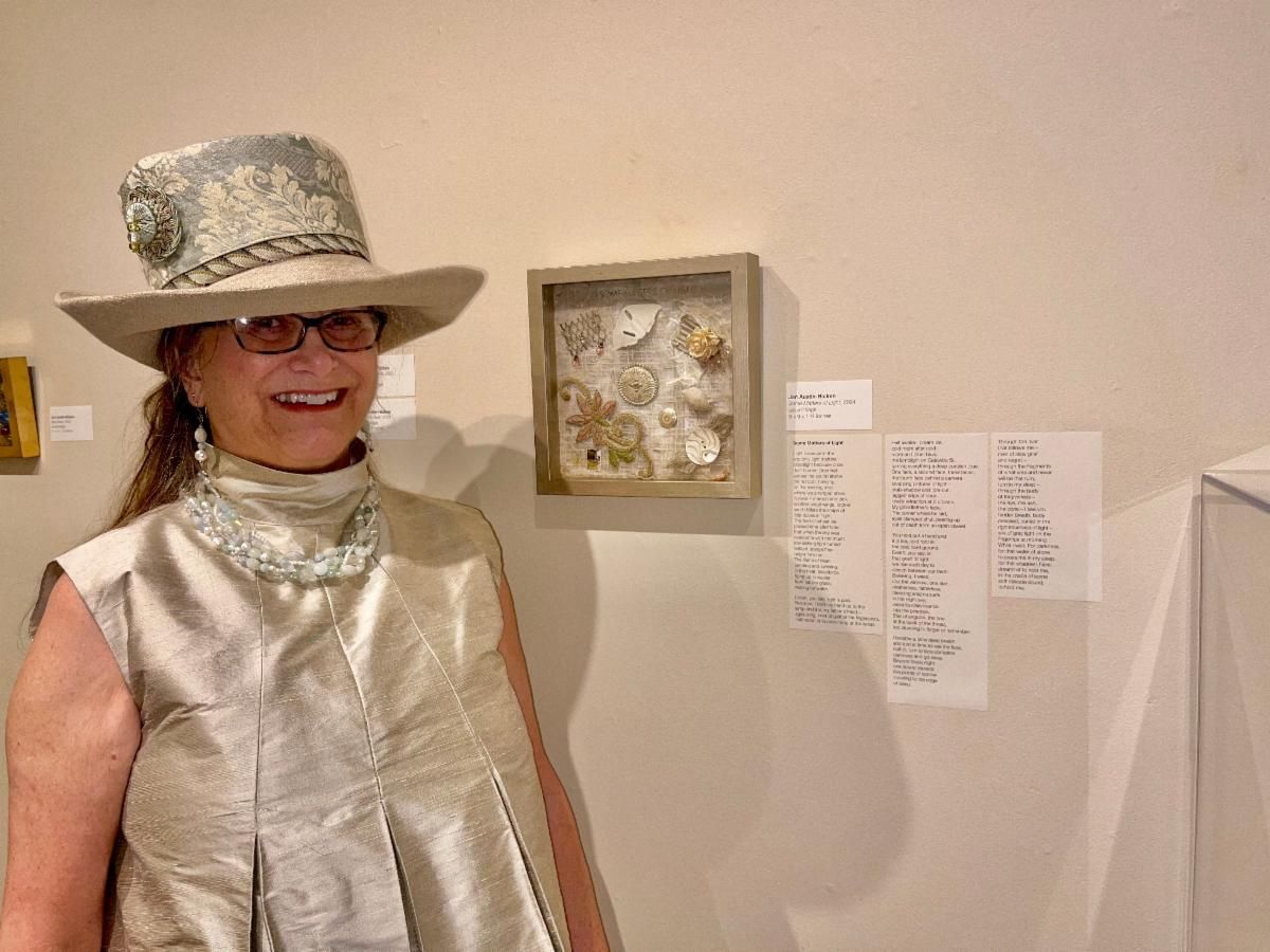 Smiling woman in a silver outfit and hat stands beside framed artwork and a wall label in a gallery