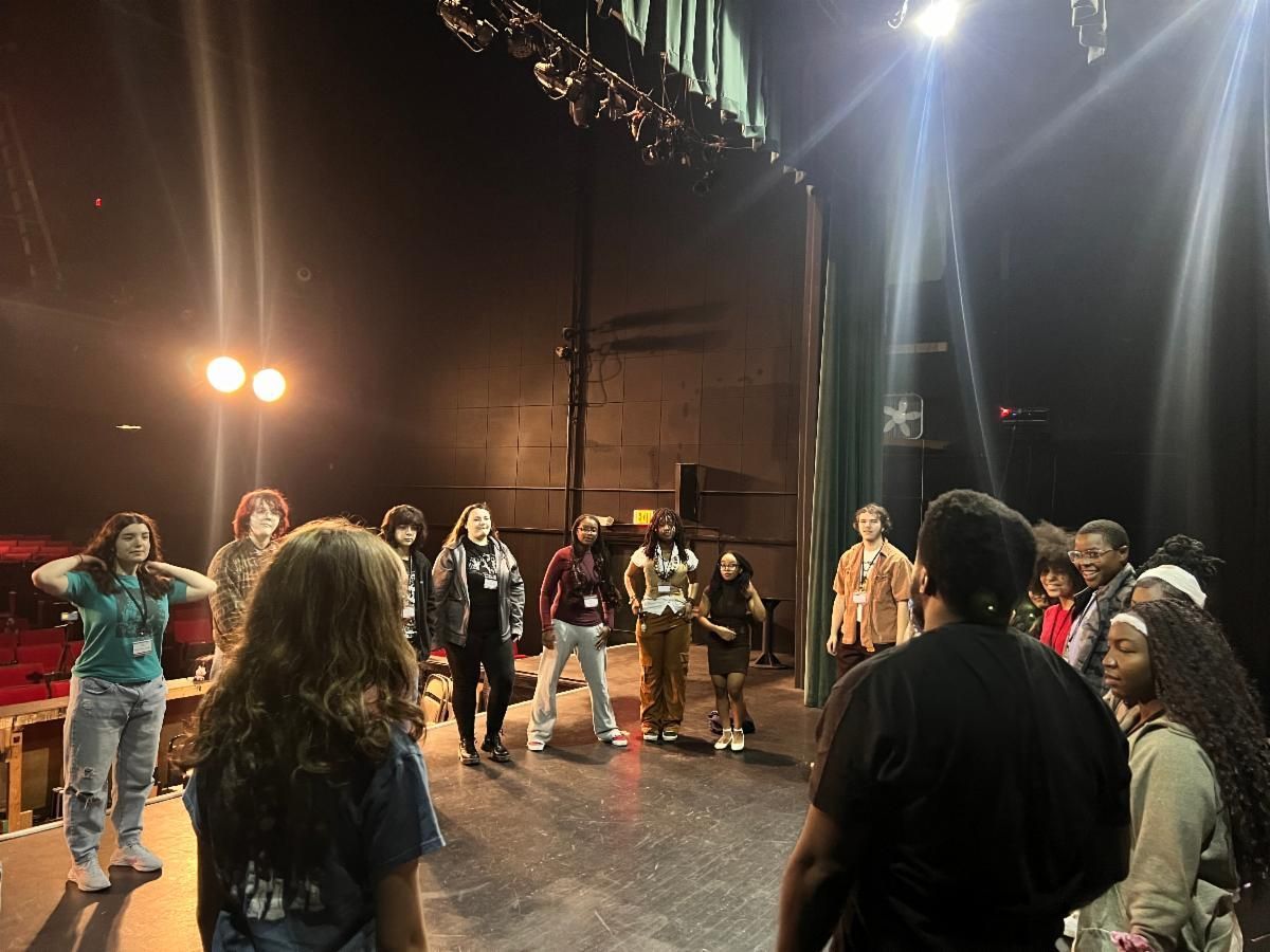 People standing in a circle on a stage under bright lights, as if in rehearsal or discussion.