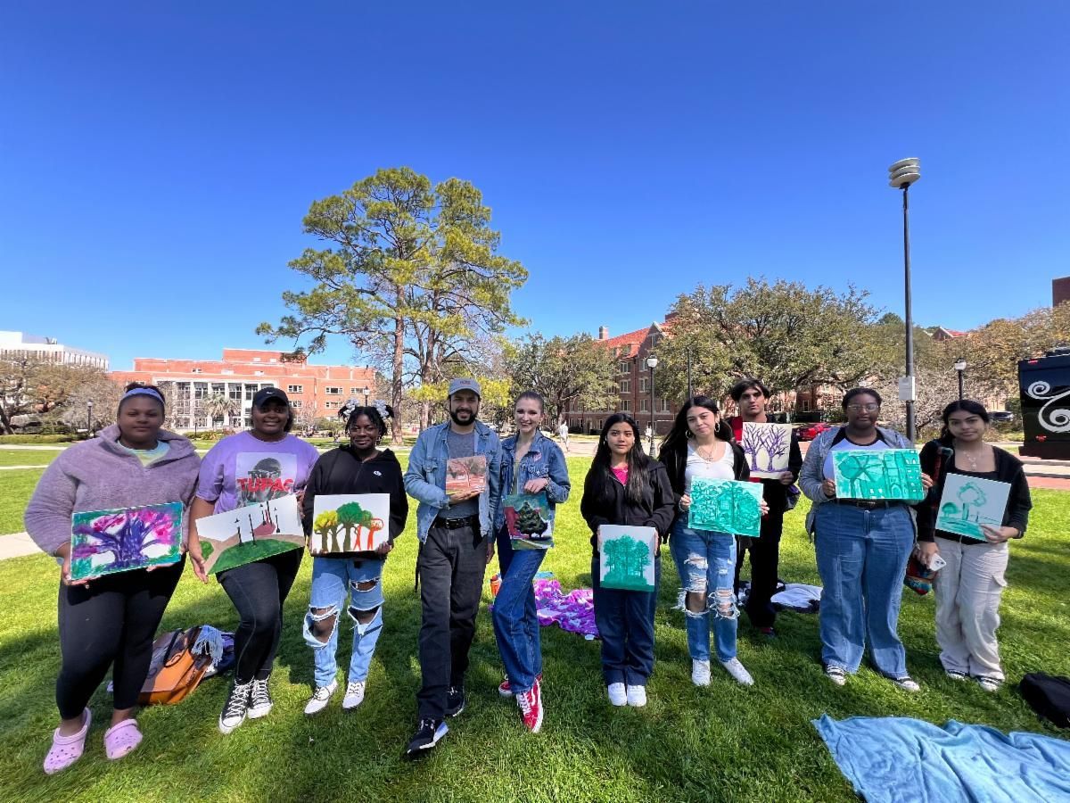 Group of people standing on a grassy lawn holding colorful paintings under a bright blue sky