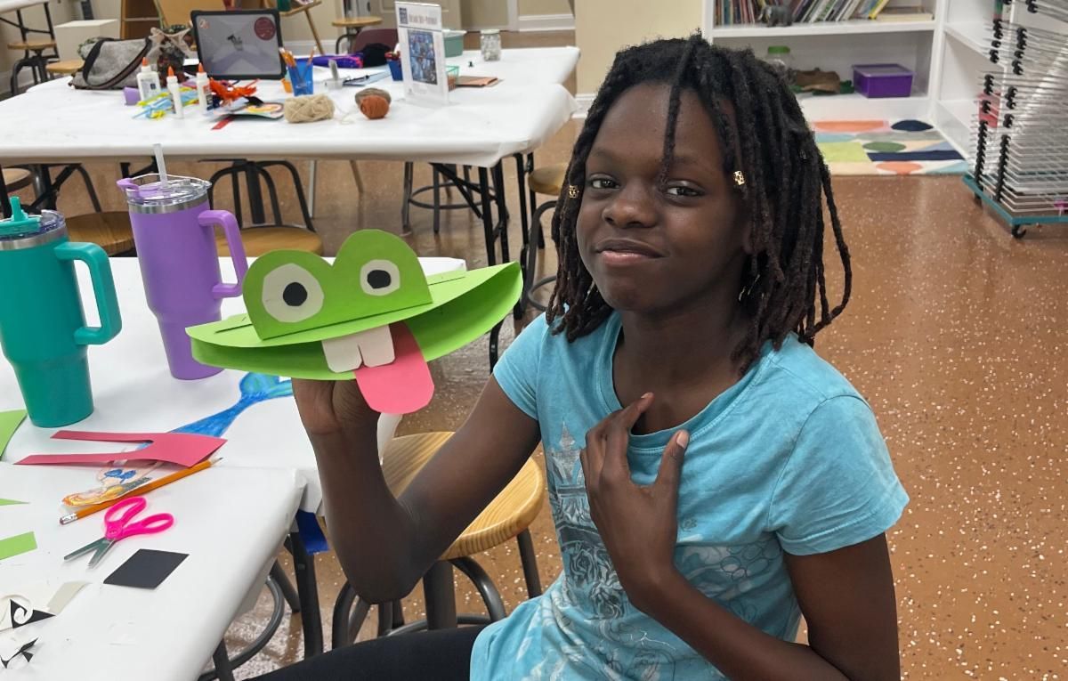 Girl holding a green craft frog at a classroom table, with supplies and tables in the background