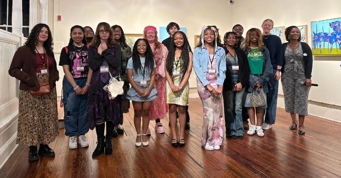 Group of people standing in a gallery, posing on a polished wooden floor with framed artwork in the background