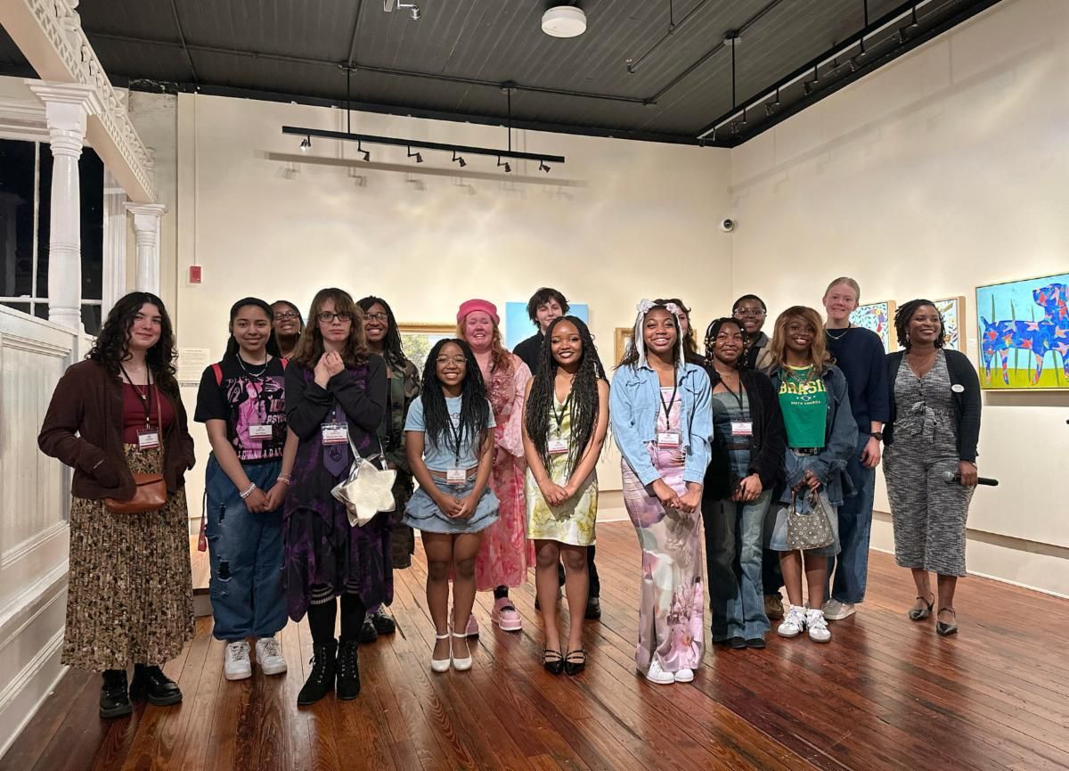 Group of people posing in an art gallery with white walls, wood floors, and framed artwork.