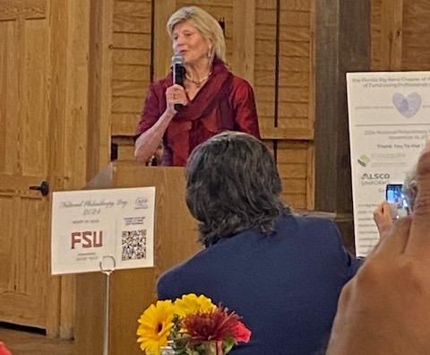 Woman speaking at a podium in a wood-paneled room, with attendees seated nearby.