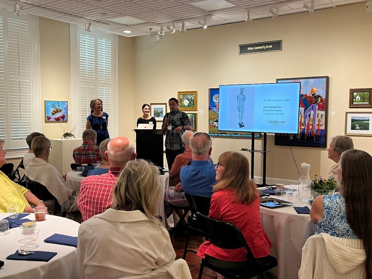 Audience watching a presentation in a gallery room, with a speaker beside a large screen and artwork on the walls