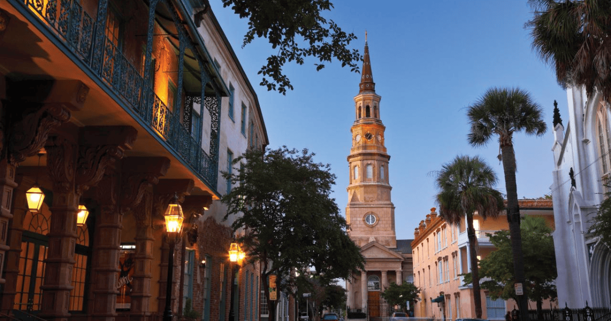 Sunset street view with a lit church tower, palm trees, and historic buildings lining the road