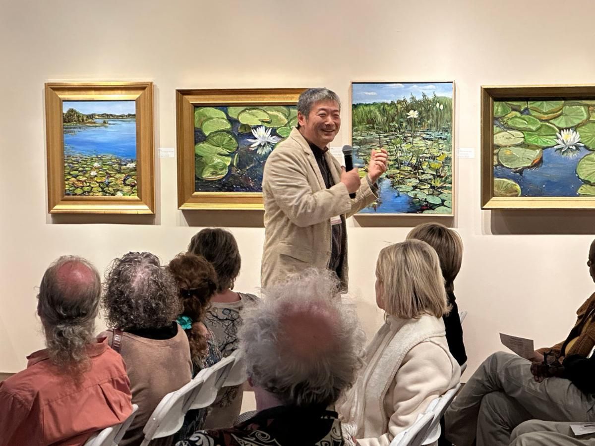 Man speaking to seated audience in an art gallery beside framed landscape paintings.