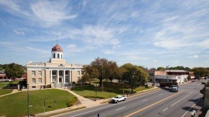 Historic courthouse with red dome beside a road, cars passing, under a partly cloudy sky.