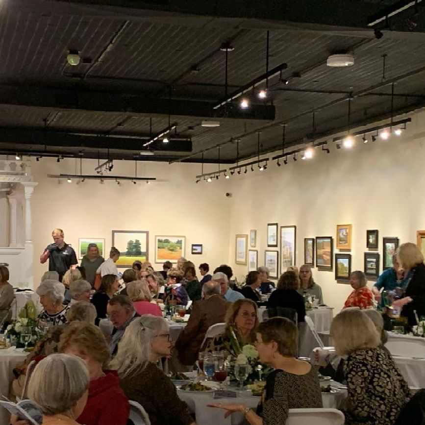 Crowded art gallery reception with seated guests, framed paintings on white walls, and track lighting overhead