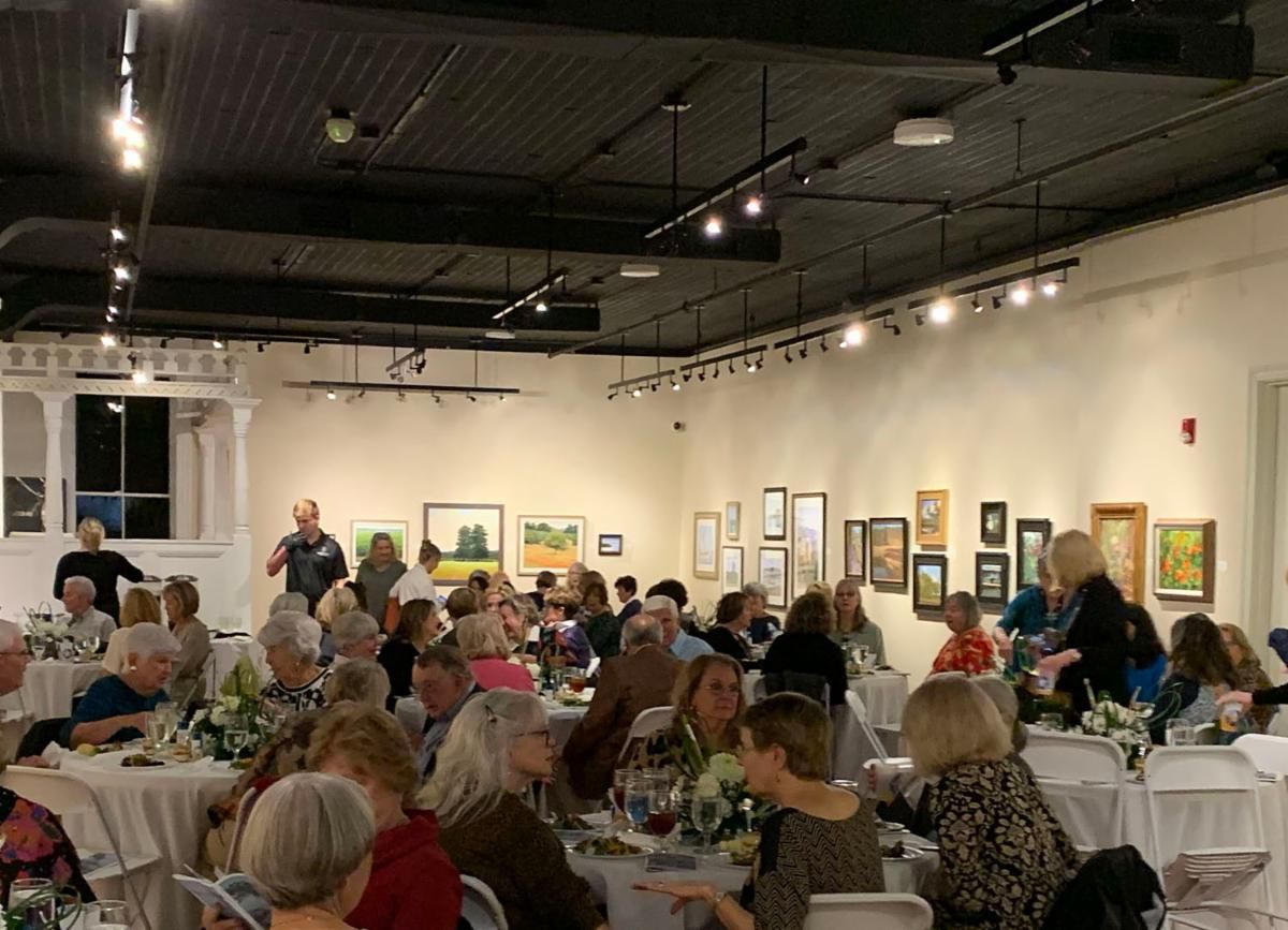 Crowded art gallery event with seated guests, framed paintings, and track lighting