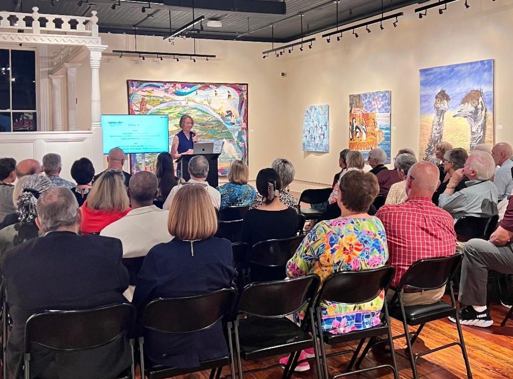 Audience seated in an art gallery listening to a speaker at a podium beside a projected slide.