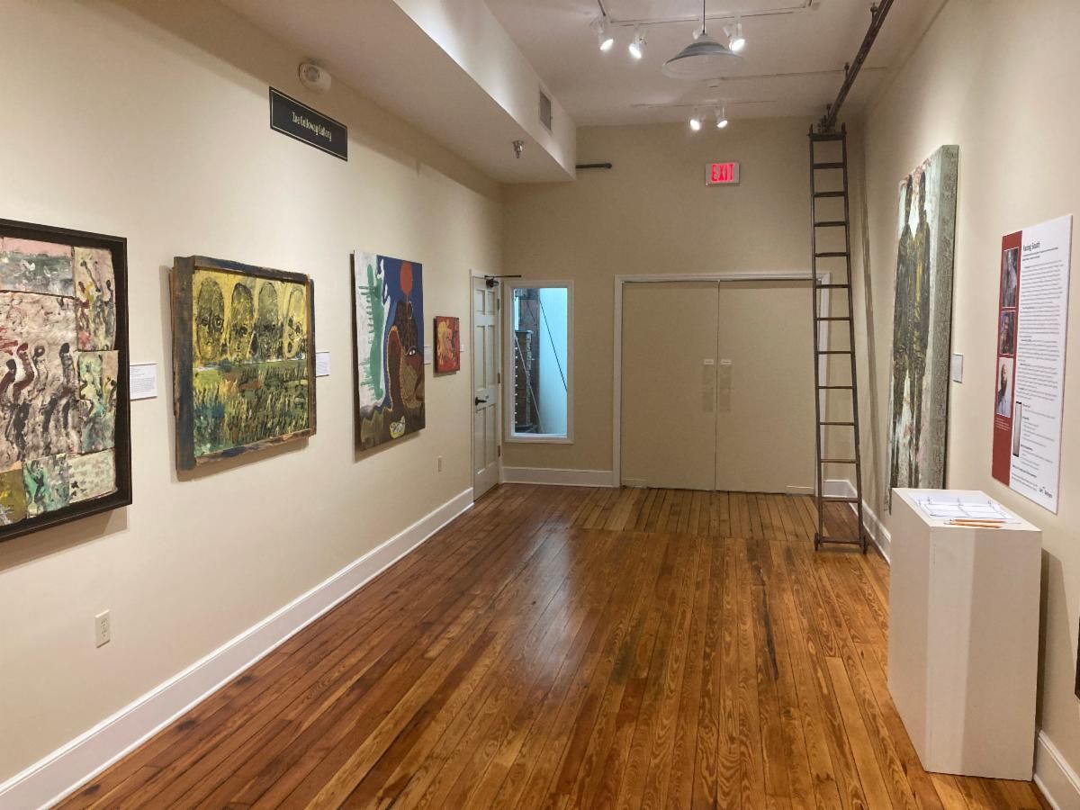 Empty art gallery with paintings on white walls and a polished wooden floor; ladder at the back.