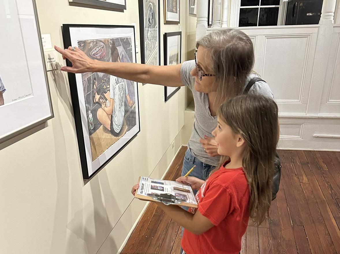 Two people in an art gallery point and examine framed artwork on a wall.