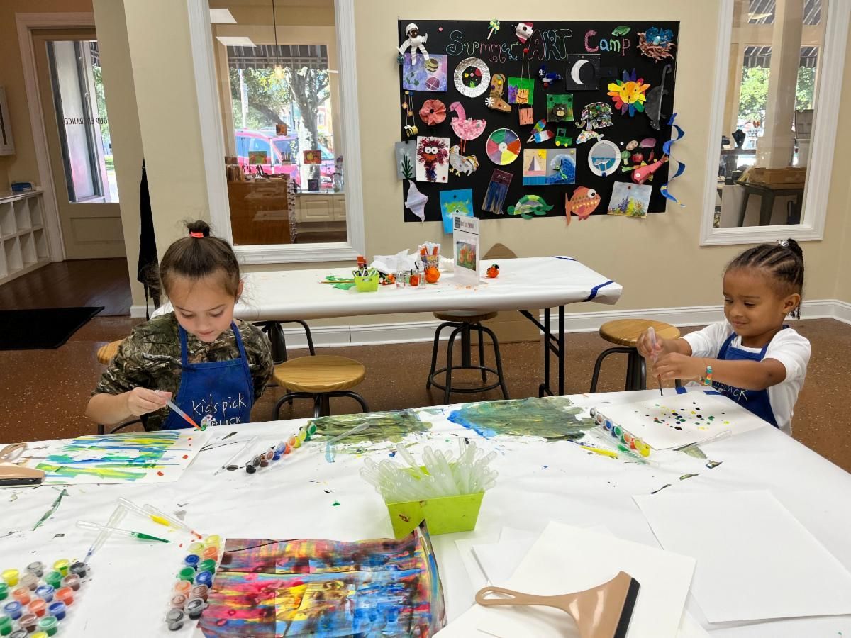 Two children painting at a craft table in a bright classroom with colorful art displayed behind them