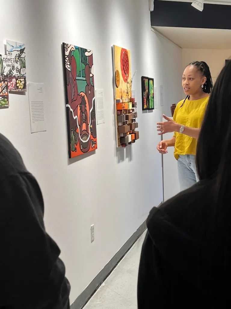 Person speaking in a gallery beside colorful wall-mounted artworks, with viewers in the foreground