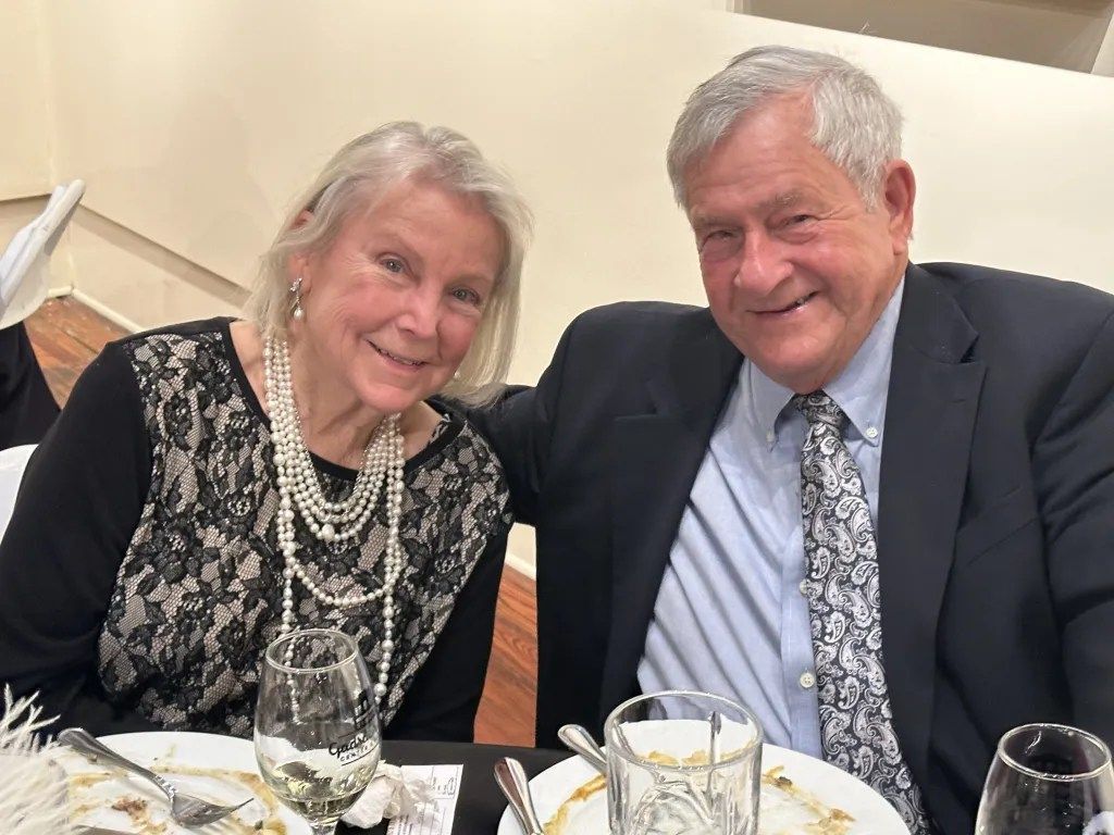 Two smiling people seated at a dinner table, dressed formally, with wine glasses and plates in front of them.