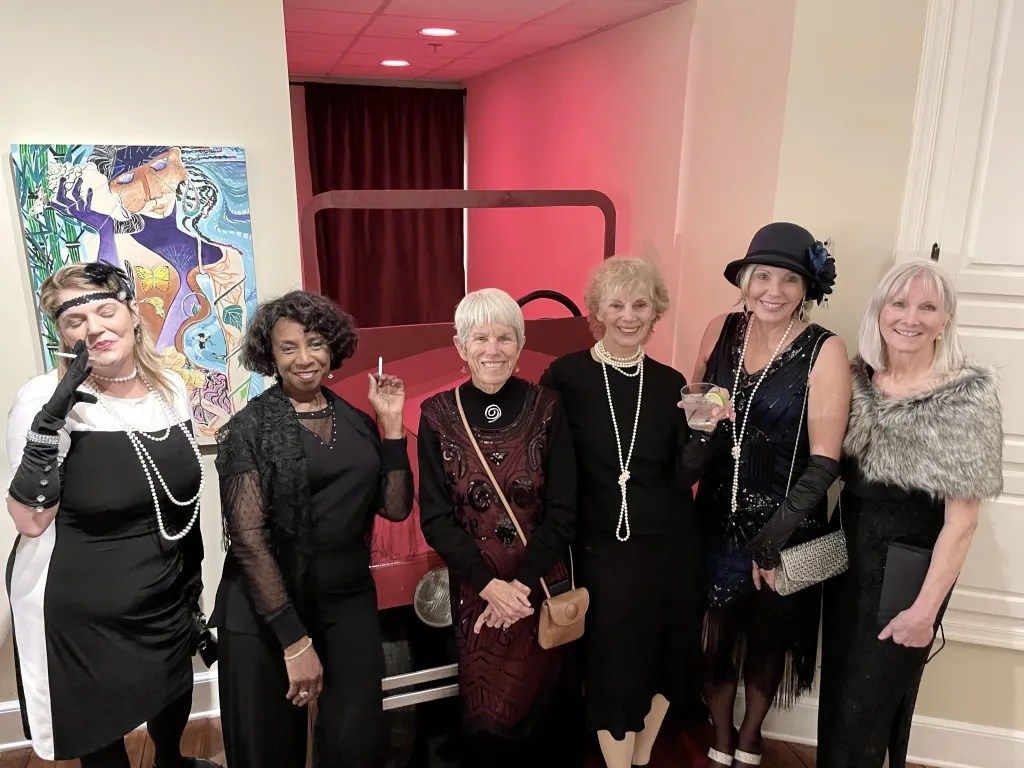 Six people posing in black outfits beside a red car in a pink room