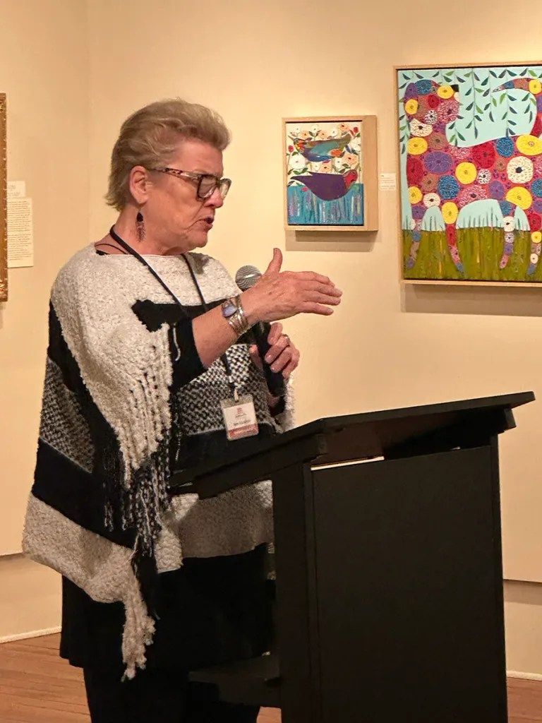 Speaker at a podium gesturing beside colorful framed artwork in a gallery