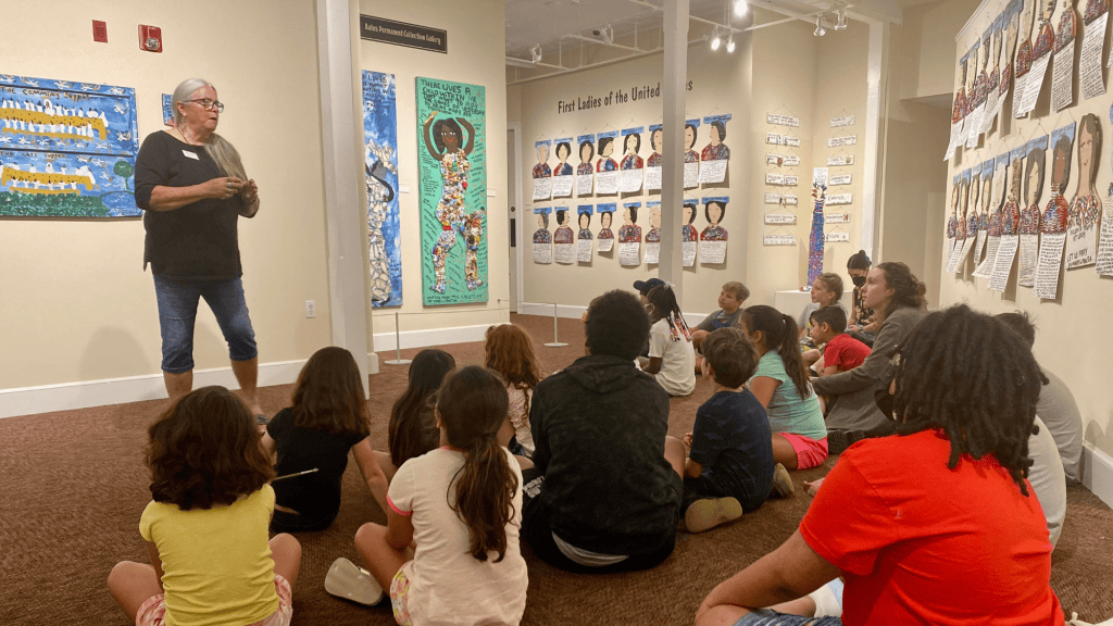 Speaker giving a presentation to seated children in an art gallery with colorful paintings on the walls