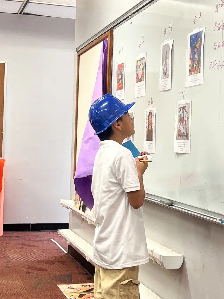 Child in a blue hard hat looking at papers on a whiteboard in a classroom or workshop setting