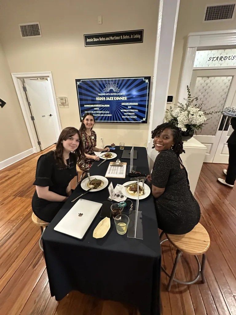 Three women seated at a table with food in a decorated indoor event space