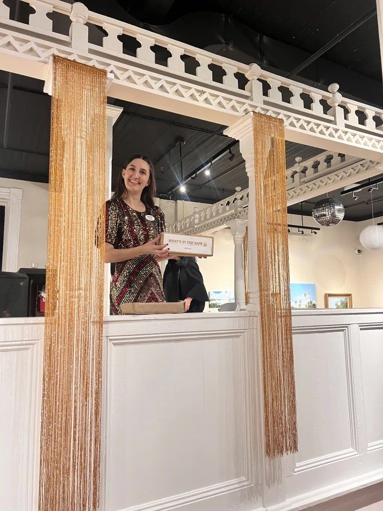 Woman standing behind a decorated white counter in a bright interior, holding a small sign and smiling.