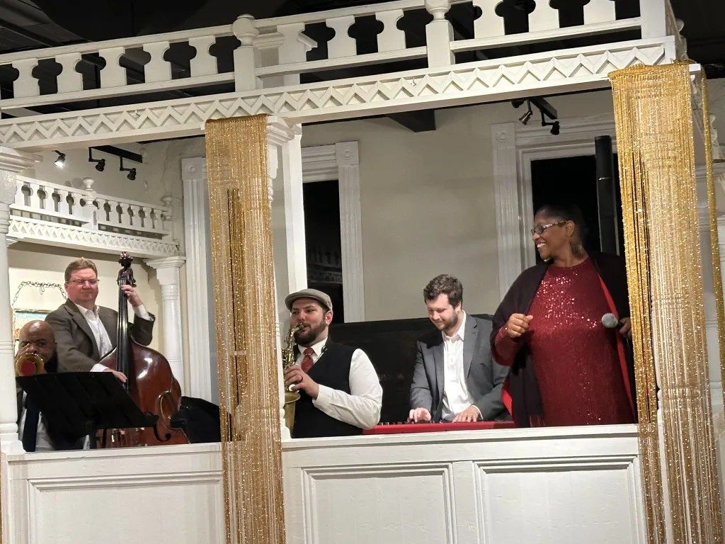 Three musicians on a balcony stage, one in a red dress, under ornate white and gold trim.