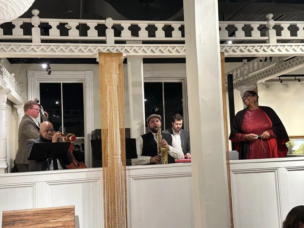 People seated at a decorated indoor event, with a man in red standing by a white railing.