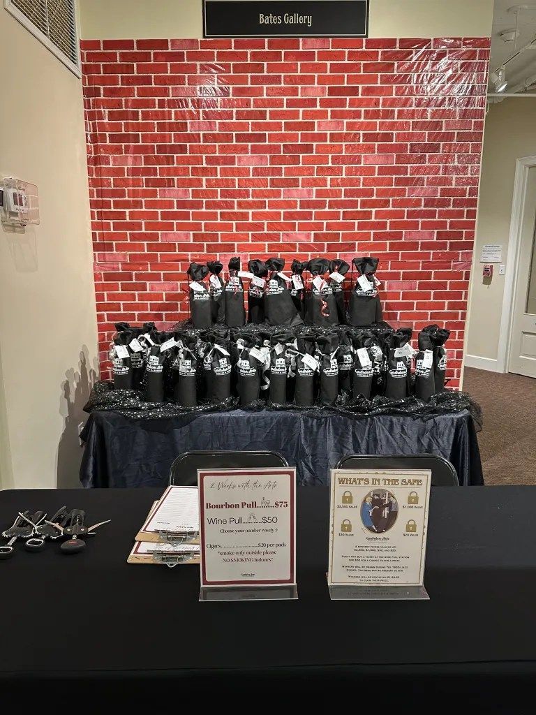 Table display of black bottles with red bows and plaques in front of a red brick wall and black tablecloth