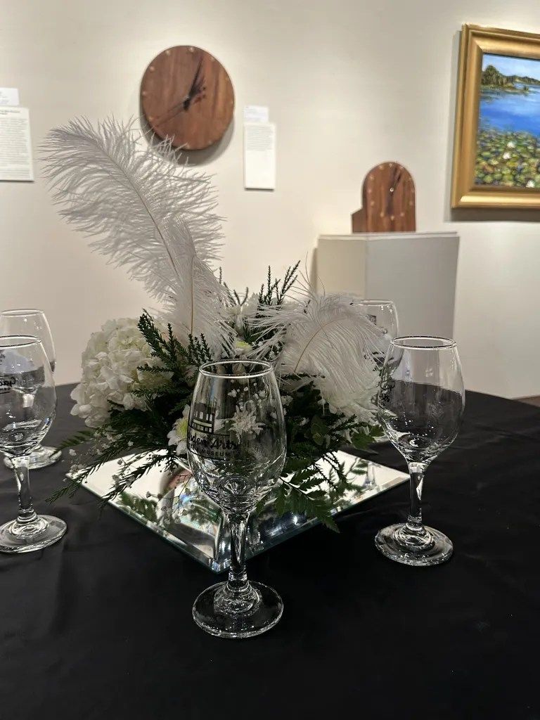 Elegant table centerpiece with white feathers, greenery, mirrored base, and wine glasses in a gallery setting
