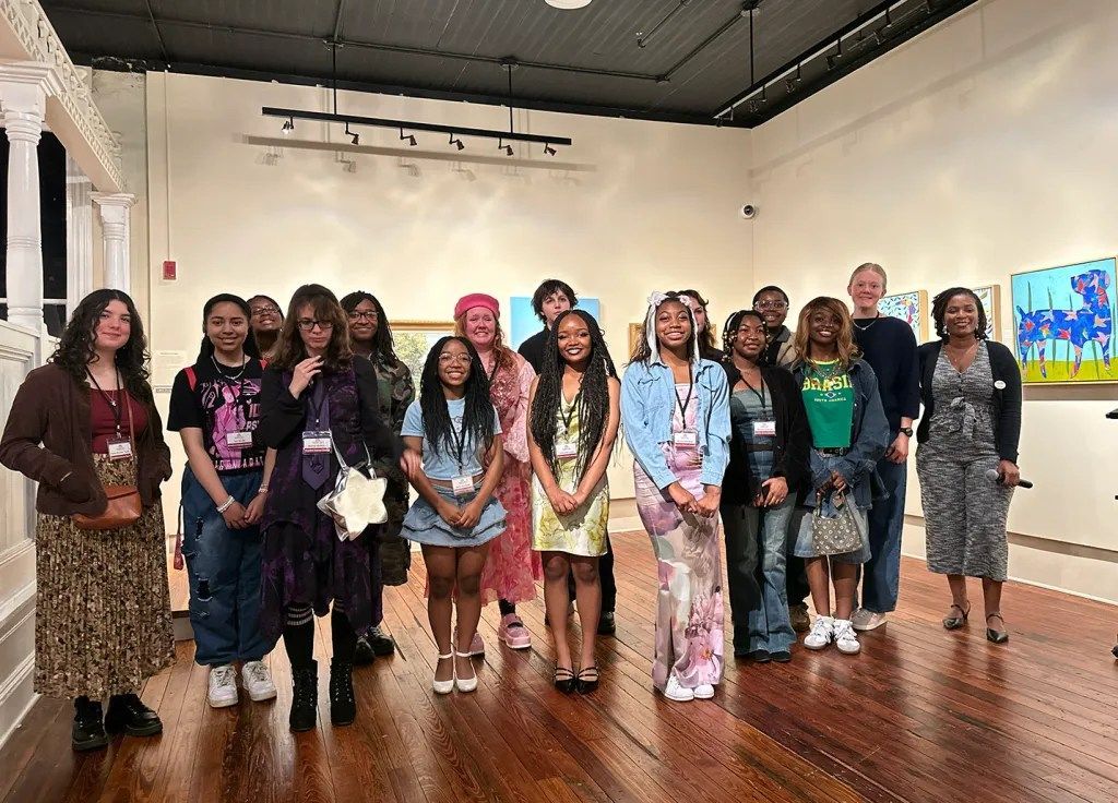 Group of people posing in an art gallery, standing on a wooden floor with framed artwork on the walls.
