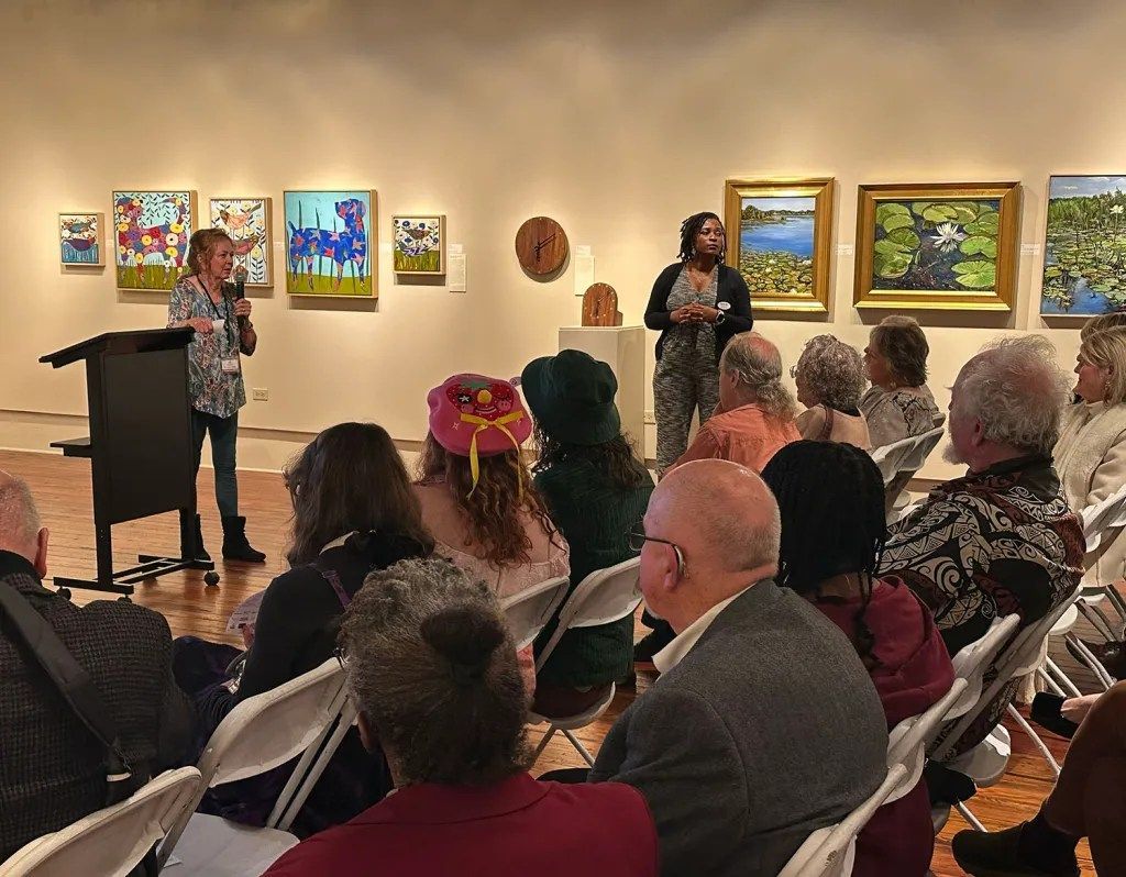 Gallery talk with two speakers addressing a seated audience in an art gallery with colorful paintings on the walls