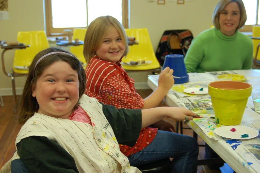 Children painting at a table in a classroom, with colorful buckets and smiling faces.