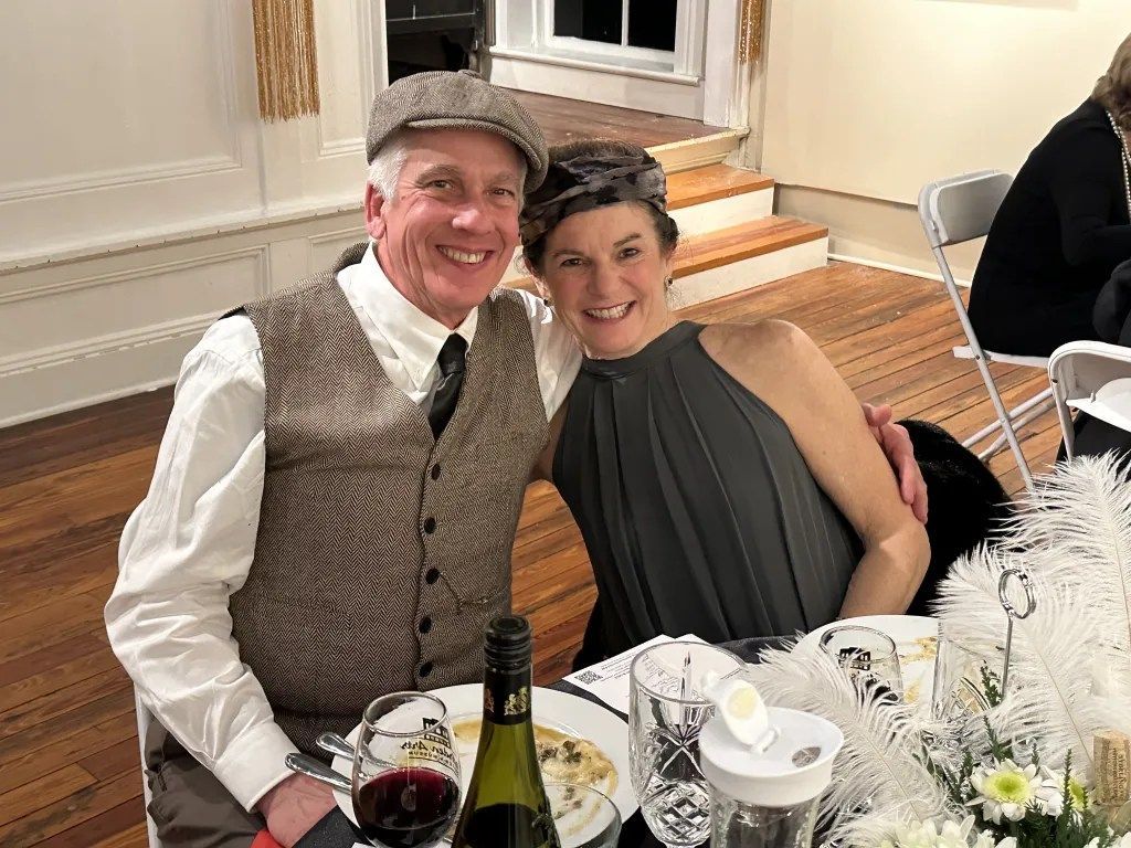 Two people smiling at a dinner table, with wine and plated food in a warmly lit room.