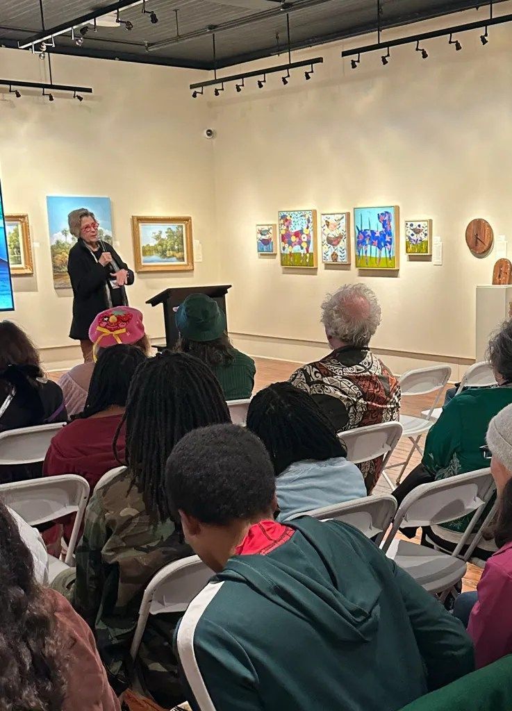Audience seated in gallery listening to a speaker beside framed artwork on white walls