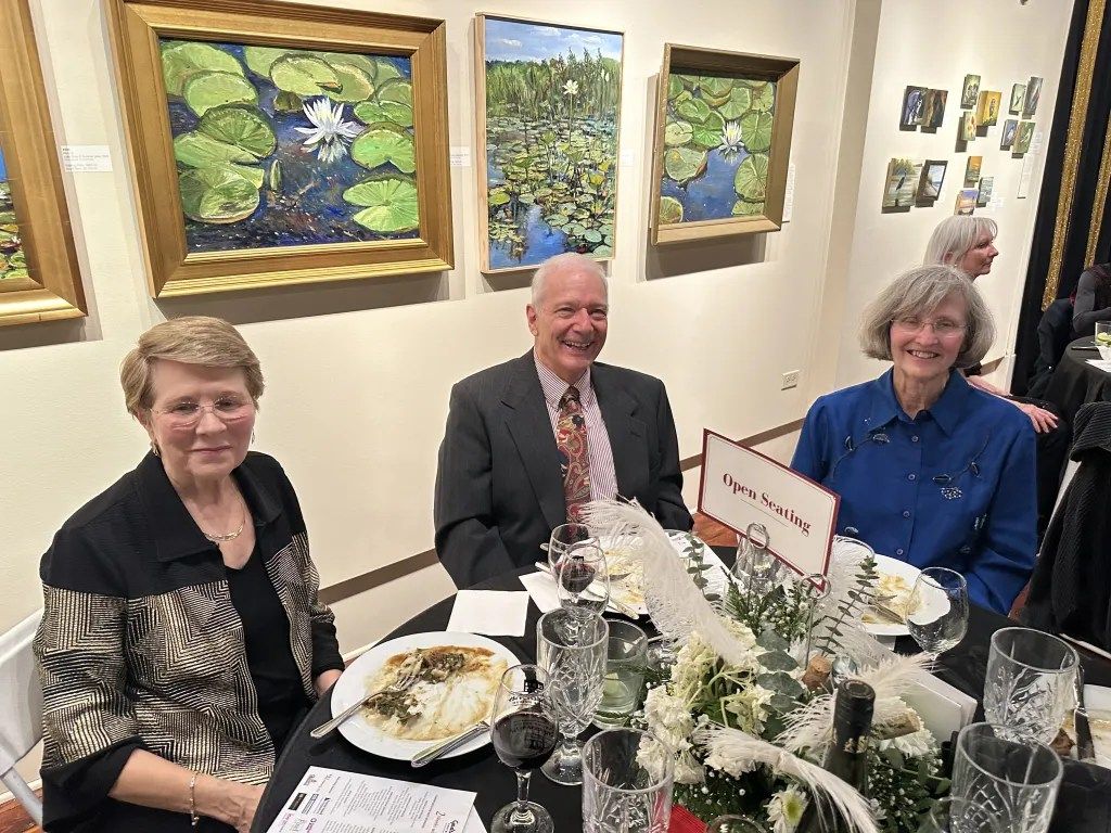Three people seated at a restaurant table with drinks and flowers, smiling near framed paintings.