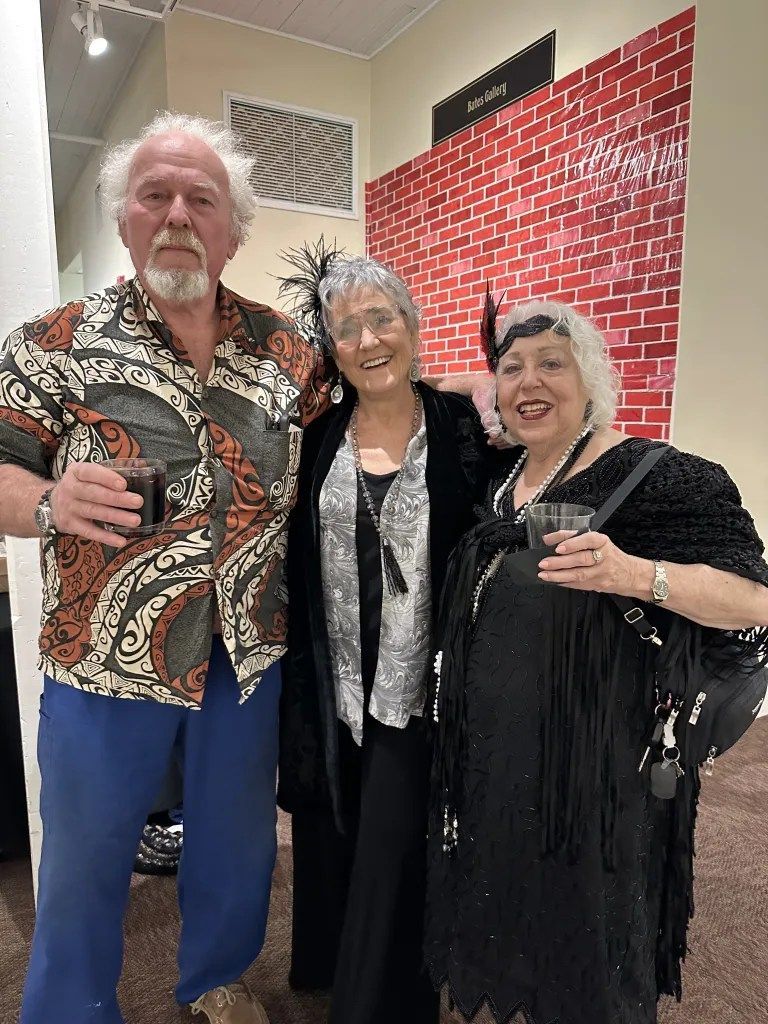 Three people posing indoors, smiling and holding drinks, with a red patterned wall behind them.