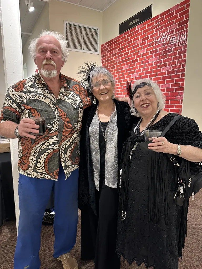 Three people standing together indoors, smiling and holding drinks, with a red brick-patterned wall behind them