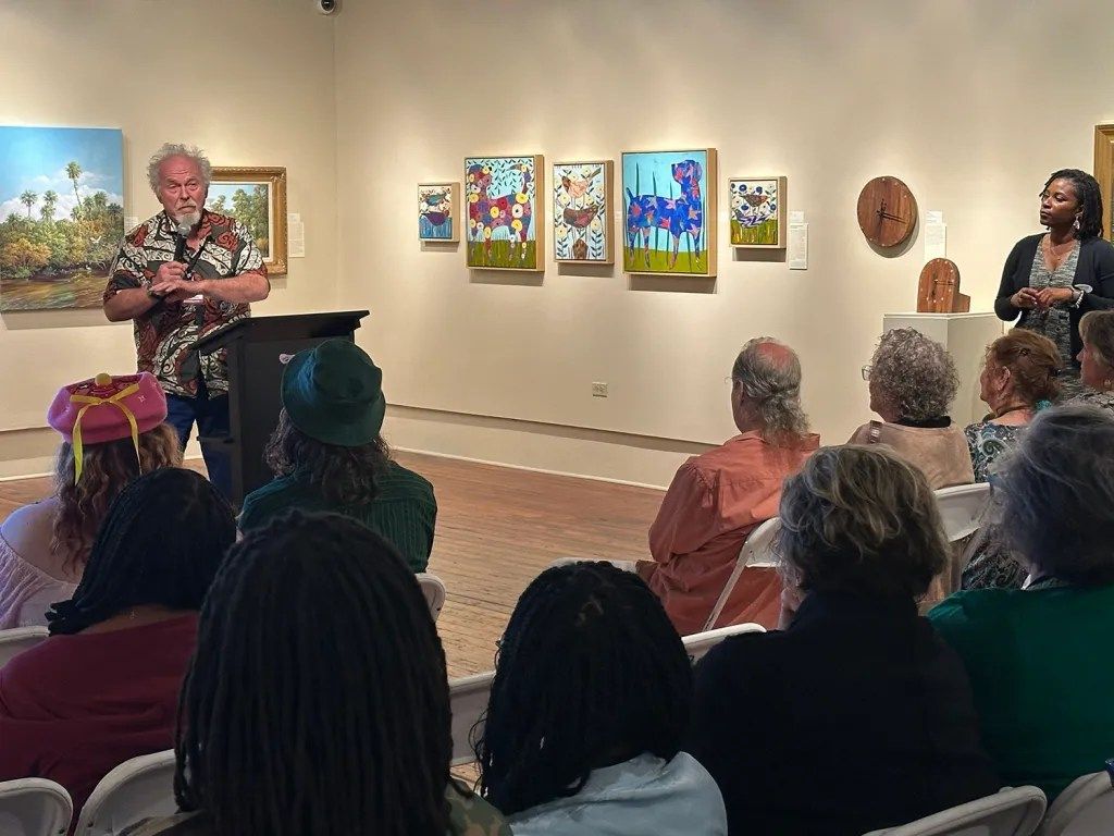 Artist speaking to seated audience in gallery with colorful paintings on the wall