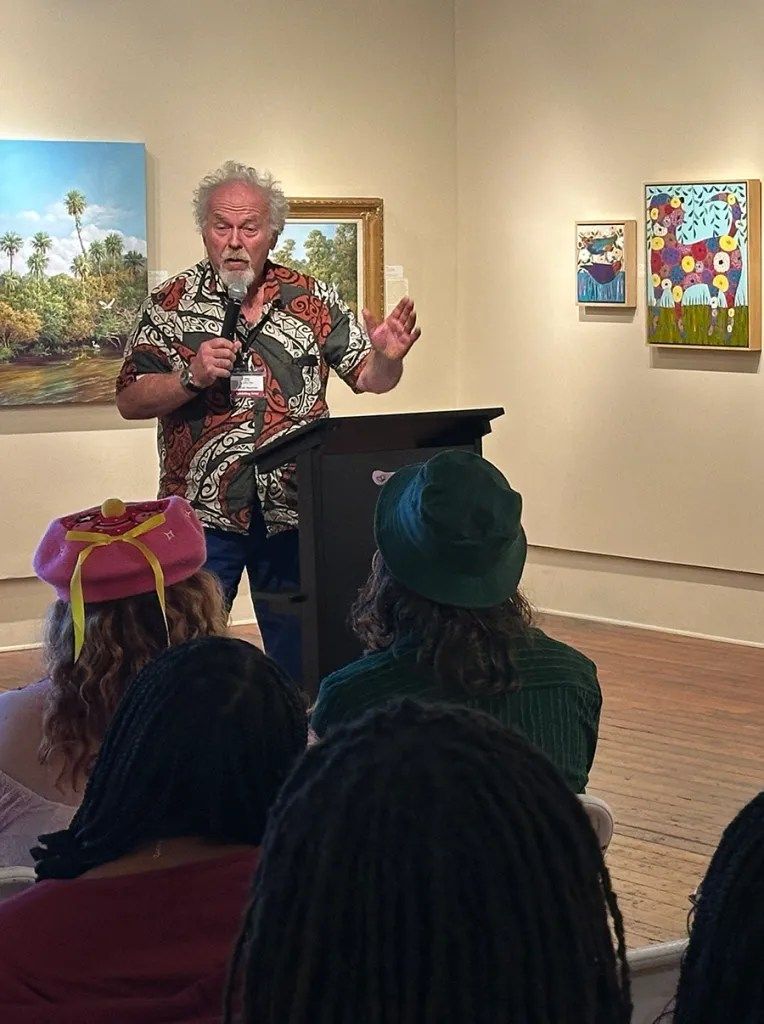 Speaker at a museum talk addressing seated audience in an art gallery, with colorful paintings on the walls.