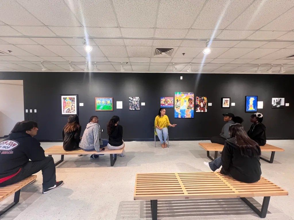 People seated on benches in a gallery, facing colorful framed artwork on a dark wall.