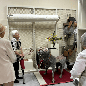Two people in a shop admire a dog display on a red rug with stuffed animals and decor on shelves.
