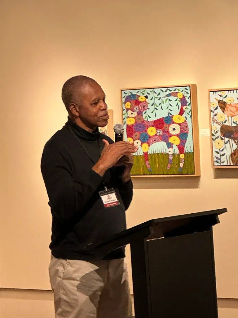 Speaker at podium in gallery, gesturing before colorful framed artwork.