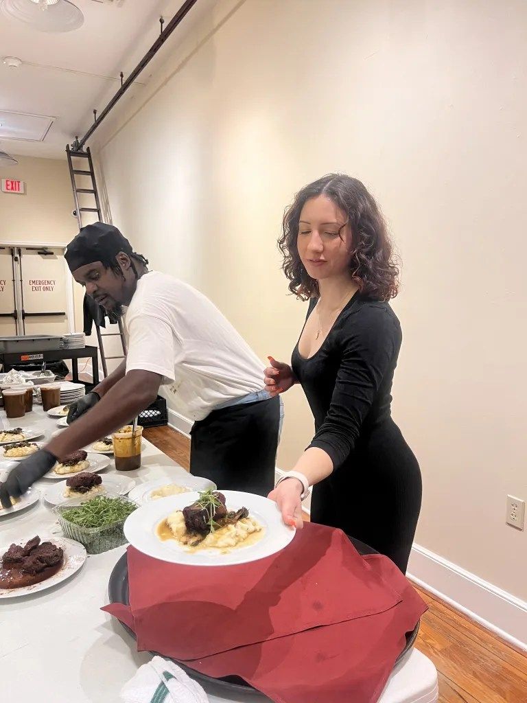People serving dishes at a buffet table in a plain indoor room, one holding a plate toward the camera.