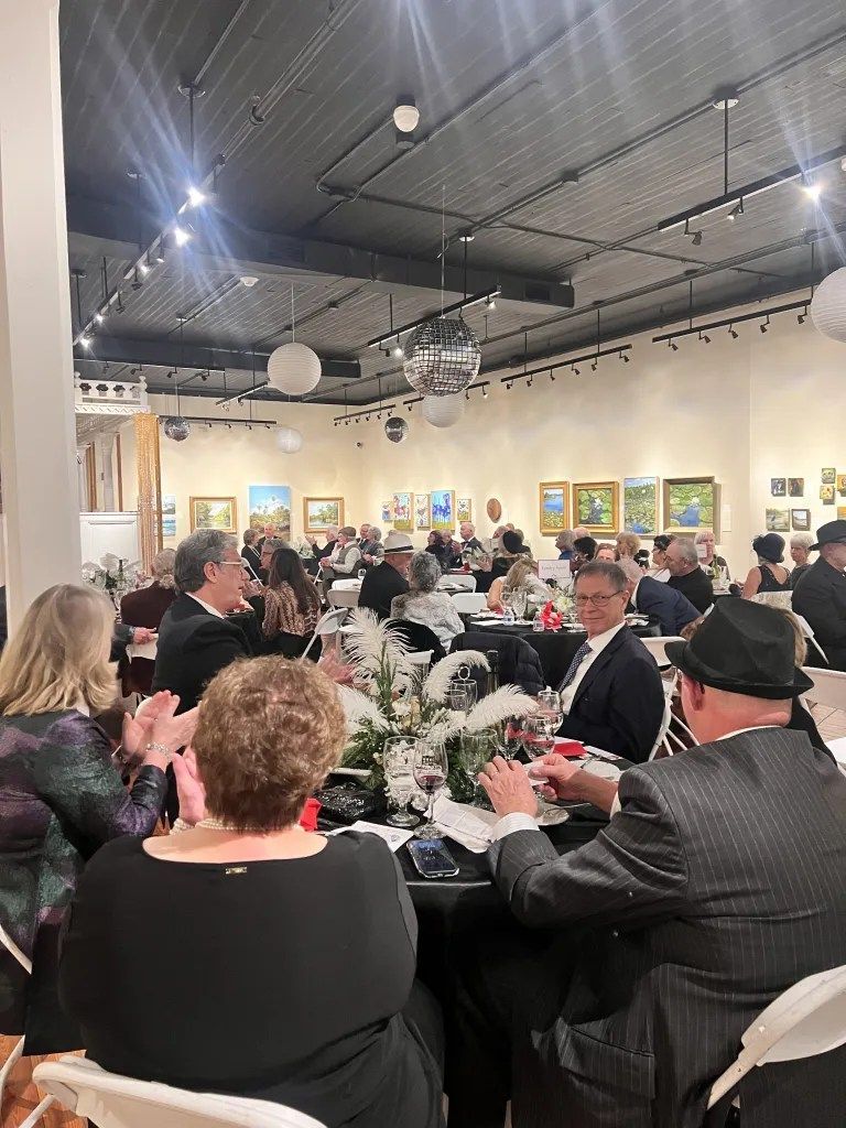 Guests dining at a formal banquet in a gallery, with tables, flowers, and framed artwork on the walls