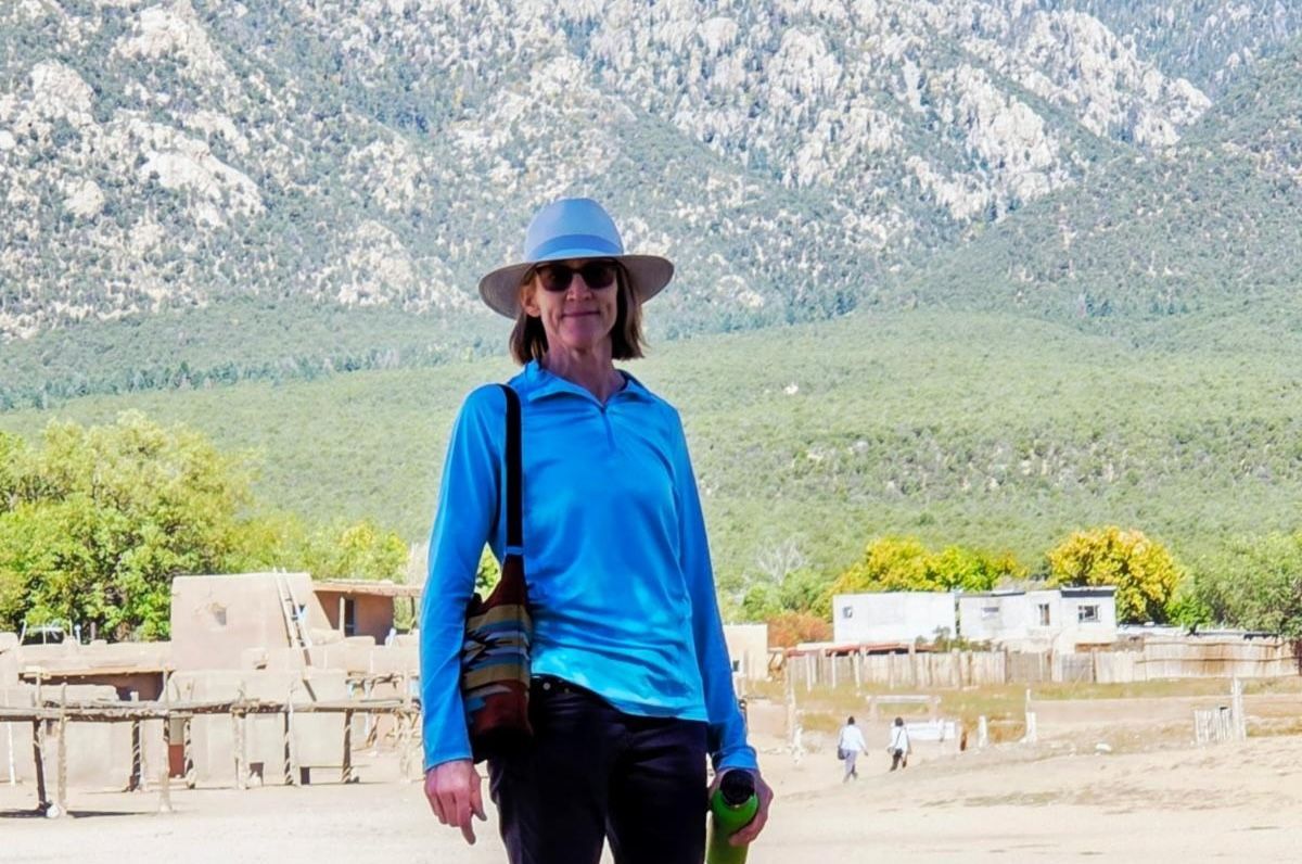 Person in blue shirt and hat standing outdoors with mountains in the background