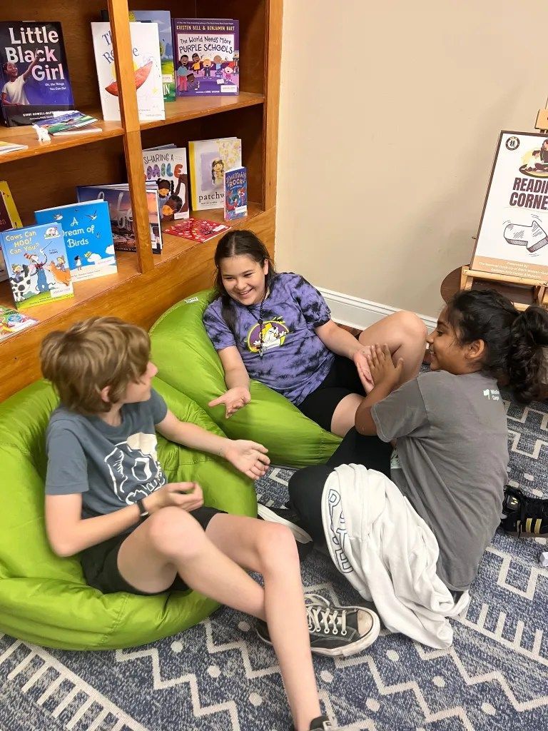 Three children sit on green beanbags, chatting in a cozy bookstore corner with shelves of books.