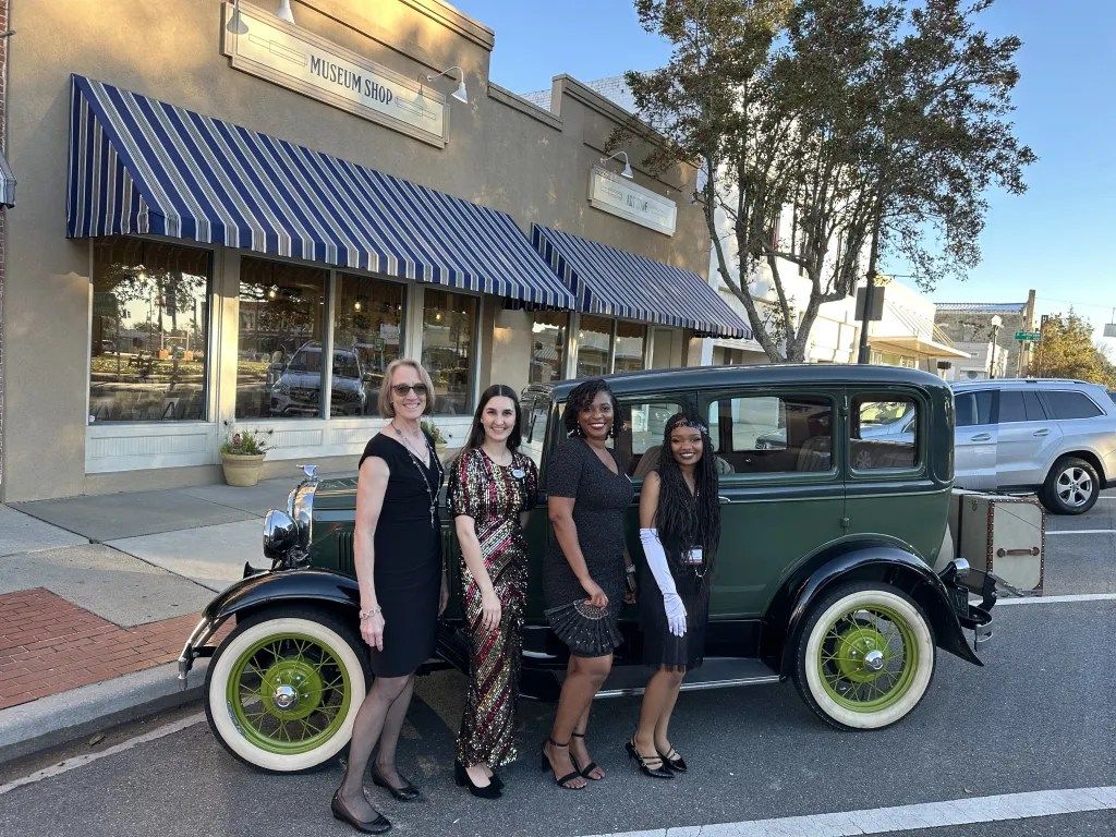 Four people posing beside a vintage green car on a downtown street outside a storefront with blue awnings