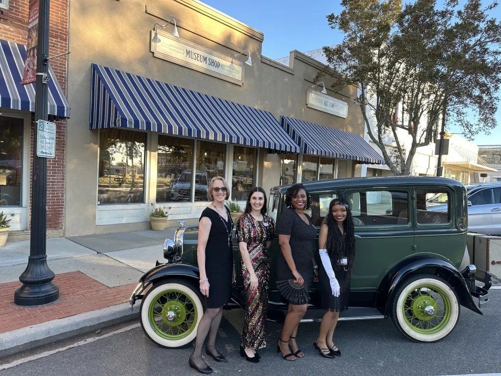 Four people stand beside a vintage green car parked on a street in front of storefronts.