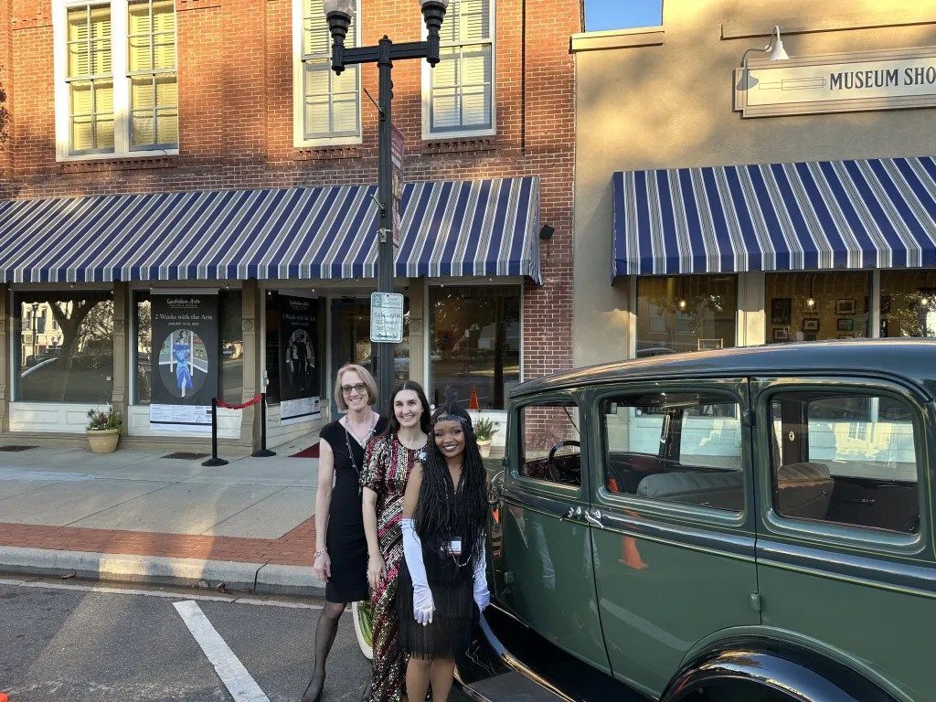 Three people standing beside a green vintage car in front of a brick storefront with striped awnings
