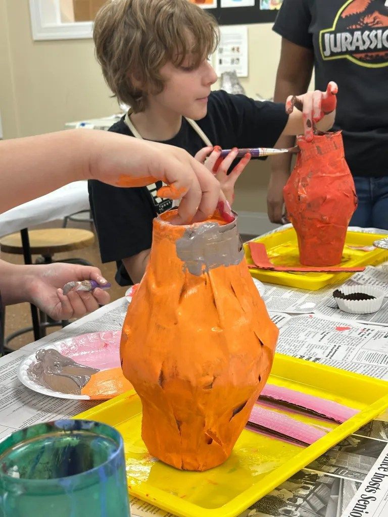 Children painting papier-mâché vases orange and red at a craft table with newspapers and paint cups